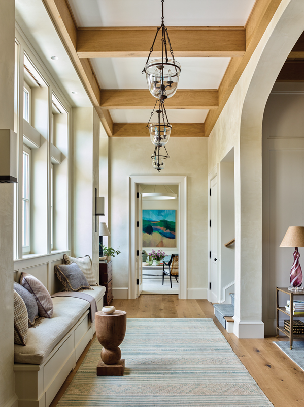 Sunshine flows in from a wall of windows above a built-in bench to the sitting room beyond. A row of glass bell jar lanterns hang over a Turkish kilim runner from Keivan Woven Arts.