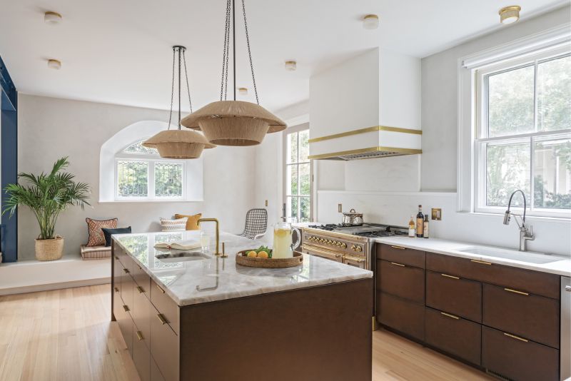 Mix It Up: A combination of metals in the kitchen, including brass and stainless steel, complement the warm, soft tones and textures of the wood cabinets and floors, string pendant lights by Nickey Kehoe, and quartzite countertops. An Italian ILVE range paired with a brass-wrapped plaster hood complete the modern yet muted tones of this now spacious, welcoming kitchen.